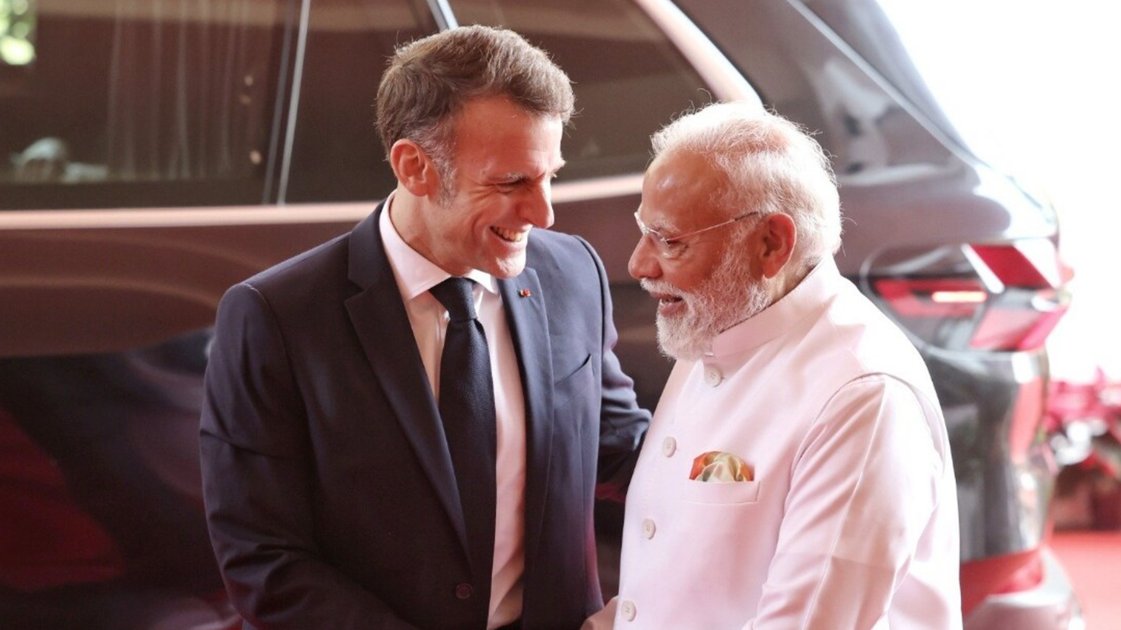 फ्रांस के राष्ट्रपति इमैनुएल मैक्रों और प्रधानमंत्री नरेंद्र मोदी हाथ मिलाते हुए, पीछे 'धुरंधर' फिल्म का पोस्टर